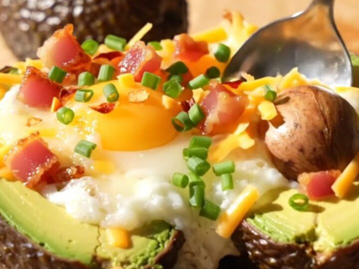 A close-up of a baked avocado half topped with a runny egg, melted cheddar, and crispy pancetta on a wooden board.