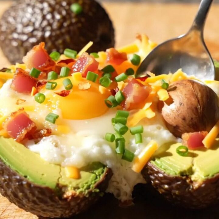 A close-up of a baked avocado half topped with a runny egg, melted cheddar, and crispy pancetta on a wooden board.