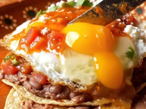 A close-up of Huevos Rancheros on a rustic clay plate with charred tortillas, fried eggs, and ranchero sauce.
