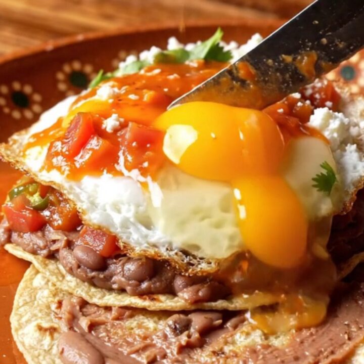 A close-up of Huevos Rancheros on a rustic clay plate with charred tortillas, fried eggs, and ranchero sauce.