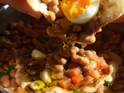 A close-up of Egyptian Ful Medames topped with halved jammy eggs, drizzled with olive oil and tahini in a rustic bowl.