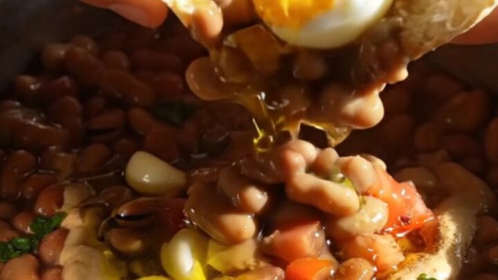 A close-up of Egyptian Ful Medames topped with halved jammy eggs, drizzled with olive oil and tahini in a rustic bowl.