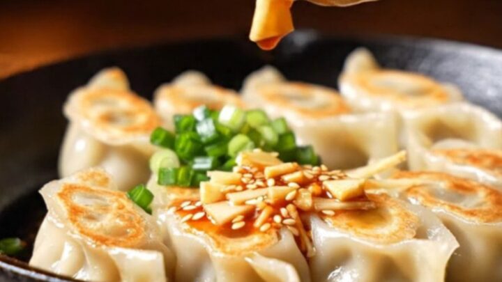 A close-up of golden-brown pork gyozas drizzled with vibrant red chili oil and sesame seeds in a dark, moody izakaya setting.