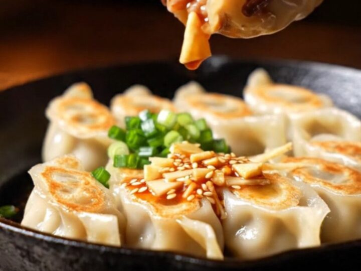 A close-up of golden-brown pork gyozas drizzled with vibrant red chili oil and sesame seeds in a dark, moody izakaya setting.