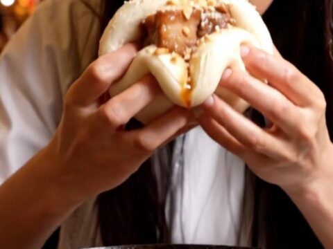 A close-up of a fluffy white steamed bao bun filled with glistening, dark amber glazed pork belly and fresh cilantro.