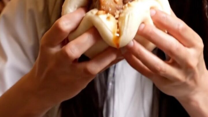 A close-up of a fluffy white steamed bao bun filled with glistening, dark amber glazed pork belly and fresh cilantro.