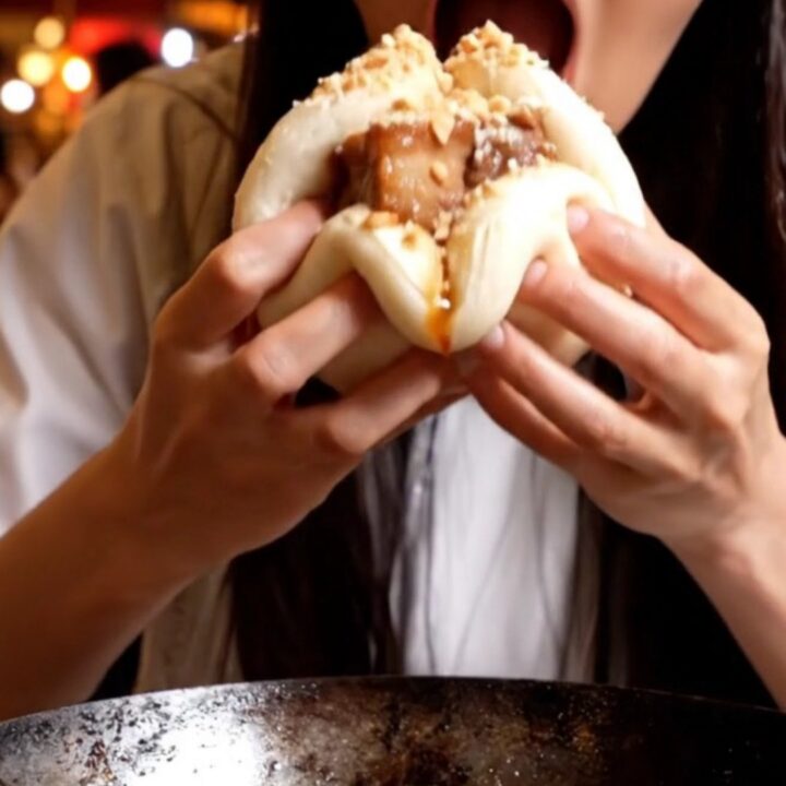 A close-up of a fluffy white steamed bao bun filled with glistening, dark amber glazed pork belly and fresh cilantro.