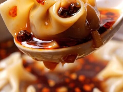 A close-up shot of silky pork wontons coated in vibrant red chili oil, garnished with crushed peanuts and green scallions.