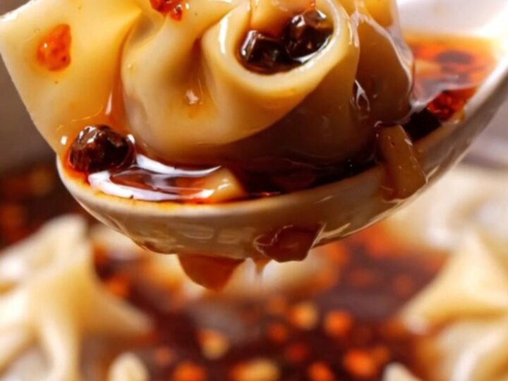A close-up shot of silky pork wontons coated in vibrant red chili oil, garnished with crushed peanuts and green scallions.