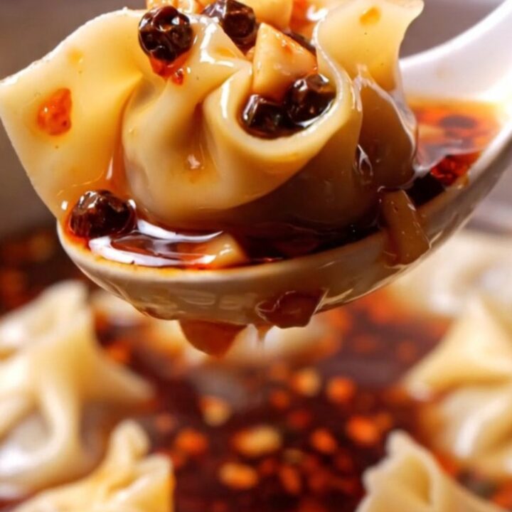 A close-up shot of silky pork wontons coated in vibrant red chili oil, garnished with crushed peanuts and green scallions.