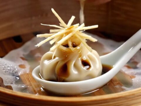 A close-up shot of a delicate 18-pleat Xiao Long Bao dumpling held by chopsticks, showing the thin, translucent skin.