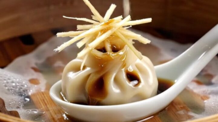 A close-up shot of a delicate 18-pleat Xiao Long Bao dumpling held by chopsticks, showing the thin, translucent skin.