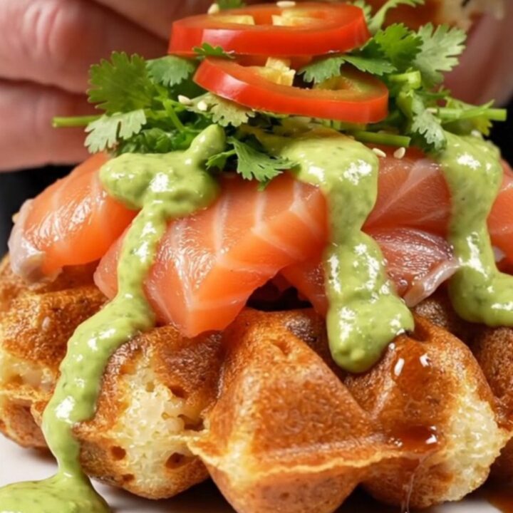 A close-up of a crispy golden sushi rice waffle topped with seared salmon belly, creamy avocado, and garnishes.