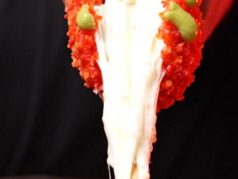 A close-up shot of a Flamin Hot Cheetos crusted Korean corn dog with a long, stretchy mozzarella cheese pull.