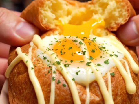 A macro shot of a fluffy, oval-shaped Korean egg bread broken in half, revealing a runny egg yolk, melted cheese, and bacon.