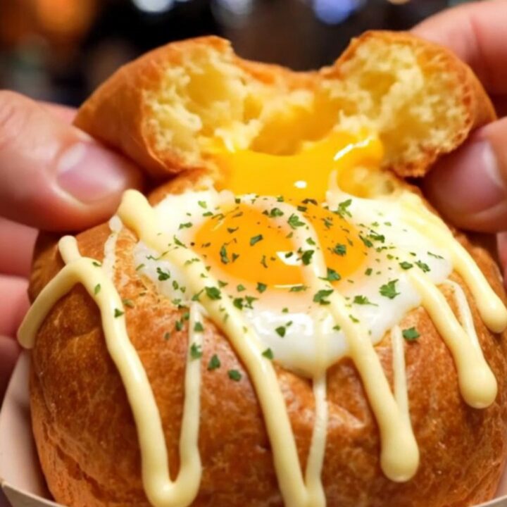 A macro shot of a fluffy, oval-shaped Korean egg bread broken in half, revealing a runny egg yolk, melted cheese, and bacon.