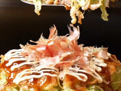 A close-up shot of a golden-brown okonomiyaki pancake topped with crispy pork belly and a feathered Kewpie mayo lattice.
