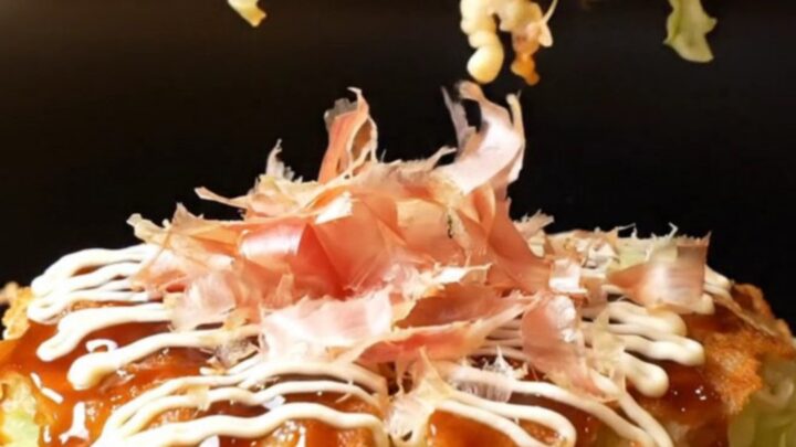 A close-up shot of a golden-brown okonomiyaki pancake topped with crispy pork belly and a feathered Kewpie mayo lattice.