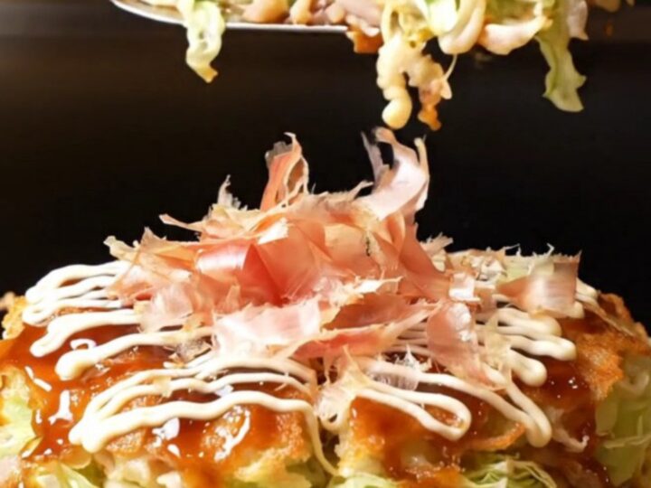 A close-up shot of a golden-brown okonomiyaki pancake topped with crispy pork belly and a feathered Kewpie mayo lattice.