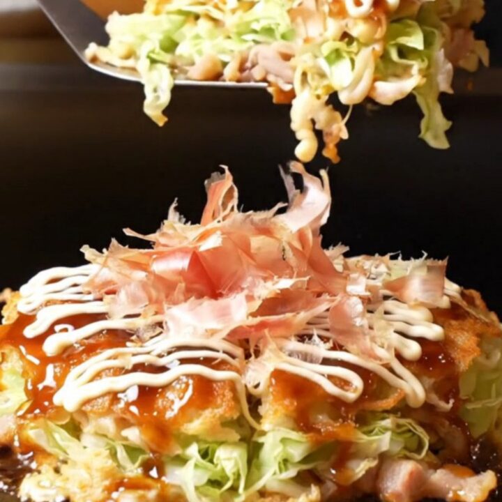A close-up shot of a golden-brown okonomiyaki pancake topped with crispy pork belly and a feathered Kewpie mayo lattice.