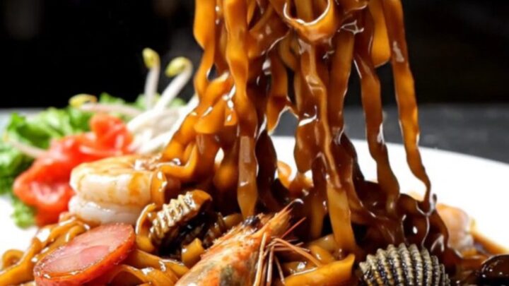 A close-up shot of smoky Penang Char Kway Teow with charred noodles, plump prawns, and sliced lap cheong sausage.