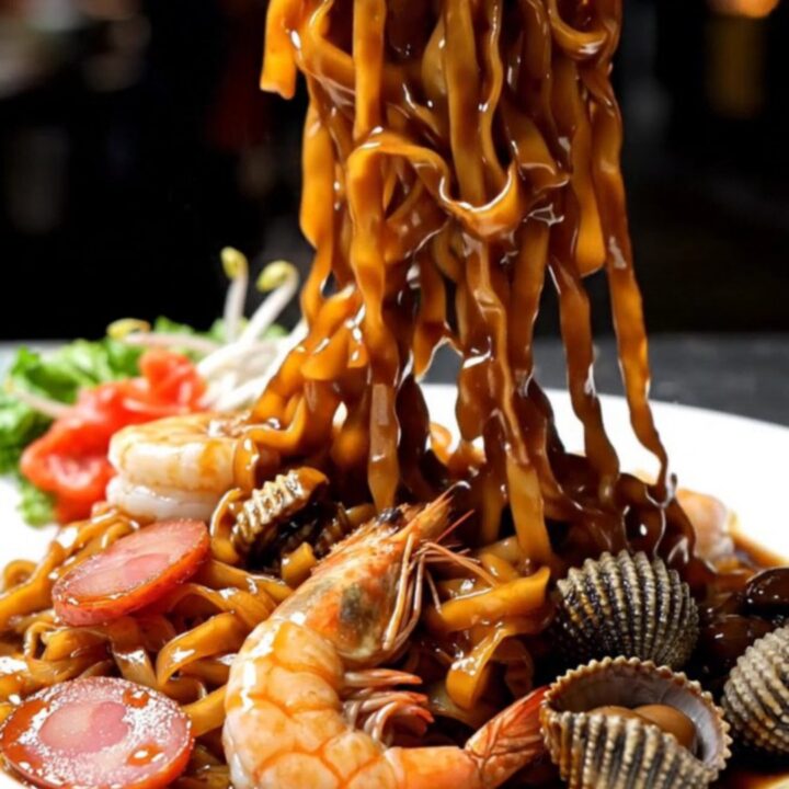 A close-up shot of smoky Penang Char Kway Teow with charred noodles, plump prawns, and sliced lap cheong sausage.