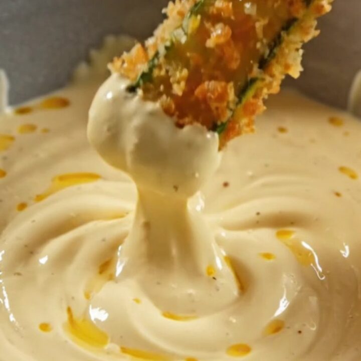A close-up of golden, crispy air-fryer zucchini fries stacked neatly next to a bowl of creamy lemon-garlic aioli.