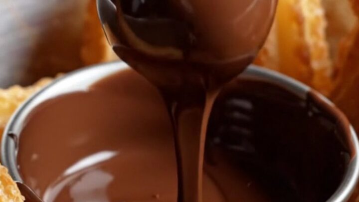 A close-up shot of a golden-brown churro bite being dipped into a bowl of rich, velvety dark chocolate ganache.