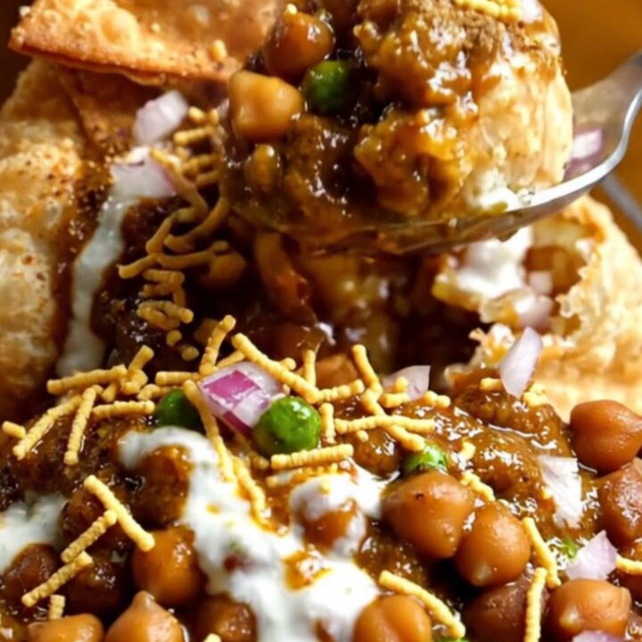 A clay bowl filled with smashed crispy samosas, spiced chickpeas, colorful chutneys, and fresh pomegranate seeds.