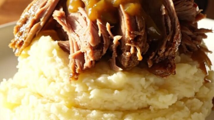 A close-up of a slow-cooked beef chuck roast being shredded with two silver forks in a black ceramic pot.