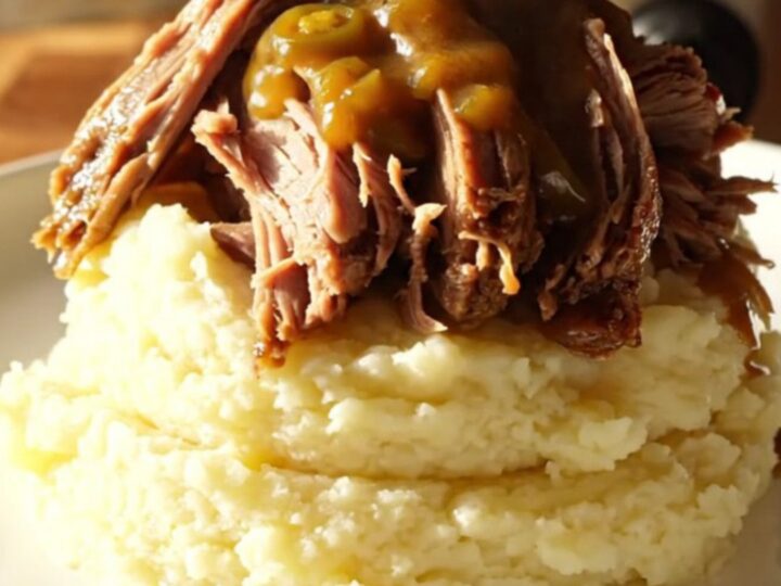 A close-up of a slow-cooked beef chuck roast being shredded with two silver forks in a black ceramic pot.