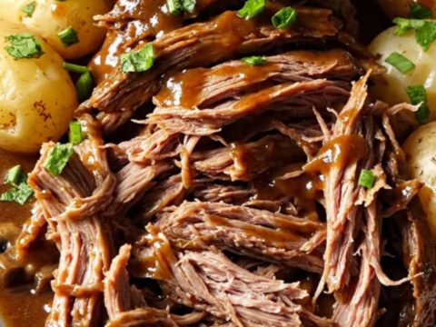 A close-up of a slow-cooked beef chuck roast in a gravy with potatoes, garnished with fresh green parsley.