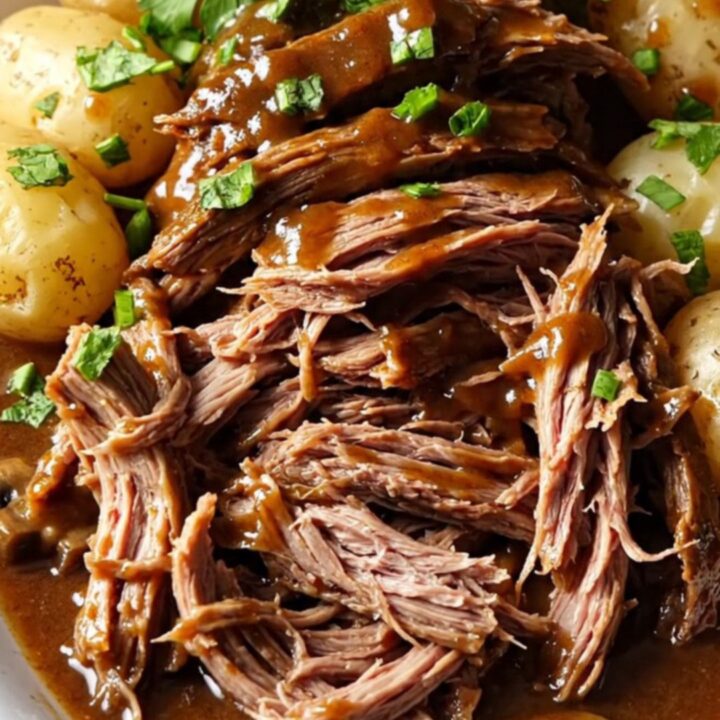 A close-up of a slow-cooked beef chuck roast in a gravy with potatoes, garnished with fresh green parsley.
