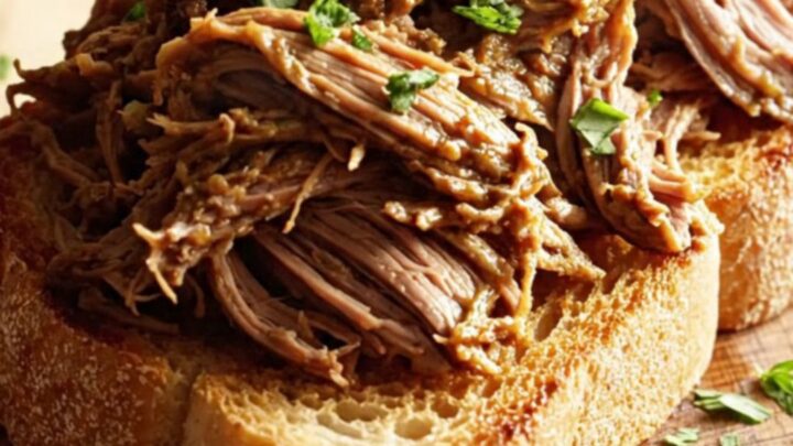 A close-up of shredded beef piled on toasted sourdough bread, drizzled with rich butter sauce and topped with fresh parsley.