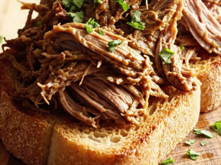 A close-up of shredded beef piled on toasted sourdough bread, drizzled with rich butter sauce and topped with fresh parsley.