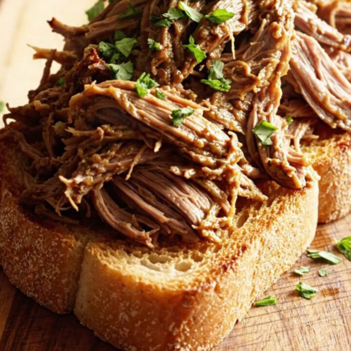 A close-up of shredded beef piled on toasted sourdough bread, drizzled with rich butter sauce and topped with fresh parsley.