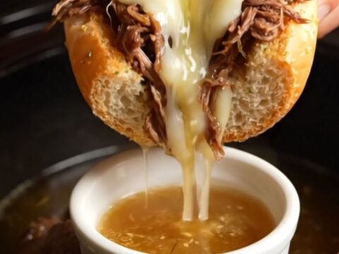 A close-up shot of a French Dip sandwich being dipped into a small ramekin of rich, dark au jus.