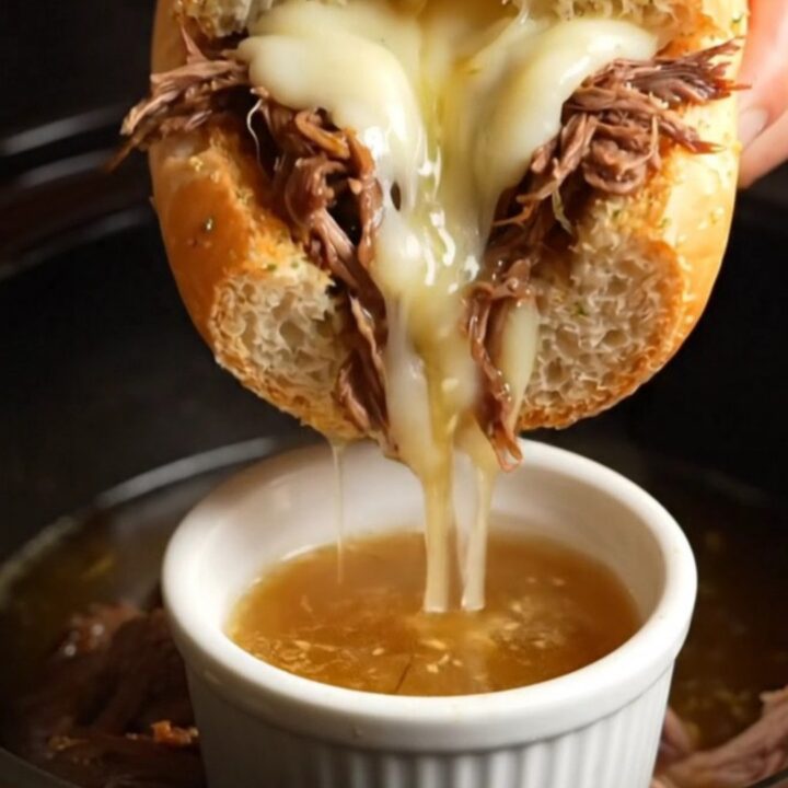 A close-up shot of a French Dip sandwich being dipped into a small ramekin of rich, dark au jus.