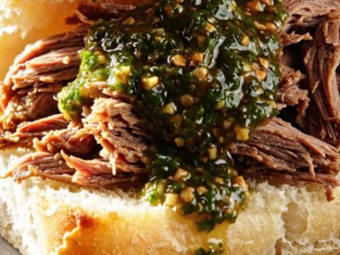 A close-up of tender, shredded beef in a ceramic slow cooker, drizzled with bright green chimichurri sauce.