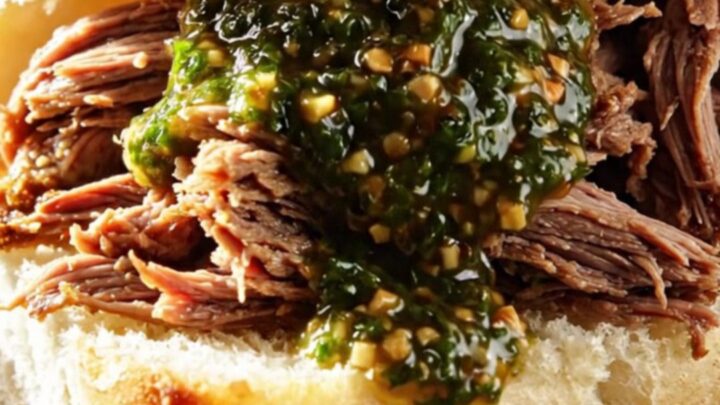 A close-up of tender, shredded beef in a ceramic slow cooker, drizzled with bright green chimichurri sauce.