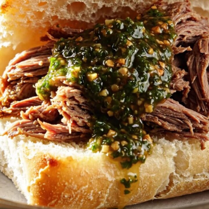 A close-up of tender, shredded beef in a ceramic slow cooker, drizzled with bright green chimichurri sauce.
