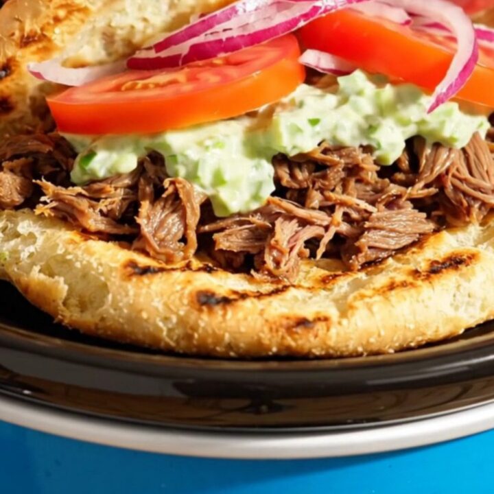 A close-up of shredded Greek beef inside a Crockpot, showing steam rising and seasoned meat.