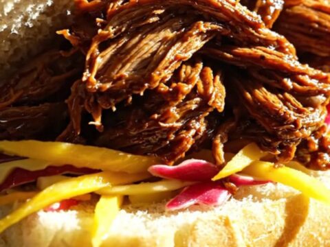 A close-up of tender, slow-cooked Jamaican jerk shredded beef served in a warm coco bread roll with fresh mango slaw.