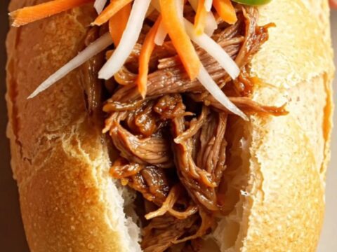 A close-up shot of a crusty Vietnamese banh mi sandwich filled with glossy, shredded lemongrass beef and fresh vegetables.