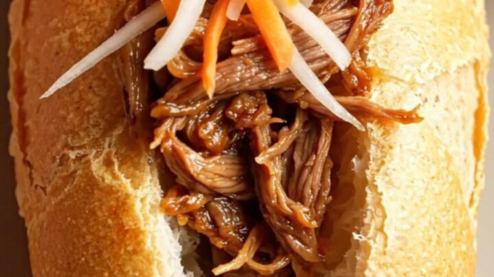 A close-up shot of a crusty Vietnamese banh mi sandwich filled with glossy, shredded lemongrass beef and fresh vegetables.