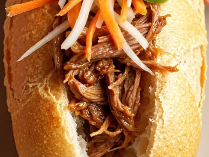 A close-up shot of a crusty Vietnamese banh mi sandwich filled with glossy, shredded lemongrass beef and fresh vegetables.