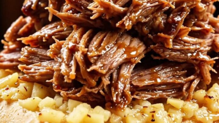A close-up of a slow cooker filled with shredded prime rib roast, garlic cloves, and rosemary in a rich butter sauce.