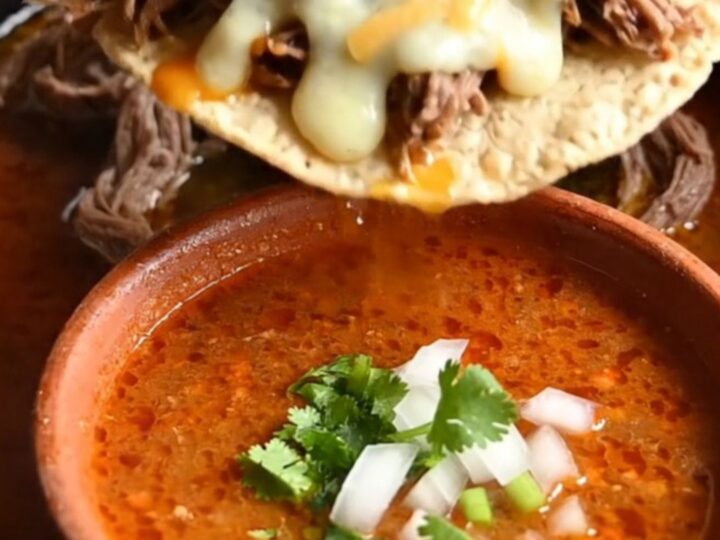 Close-up of a crispy quesataco being dipped into a bowl of rich, red birria consommé.