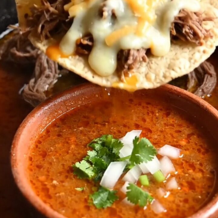 Close-up of a crispy quesataco being dipped into a bowl of rich, red birria consommé.