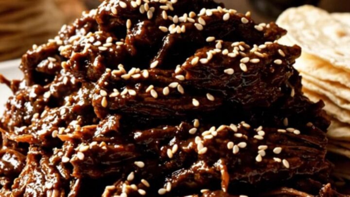 A close-up shot of shredded beef coated in a dark, glossy mole sauce, topped with toasted sesame seeds.